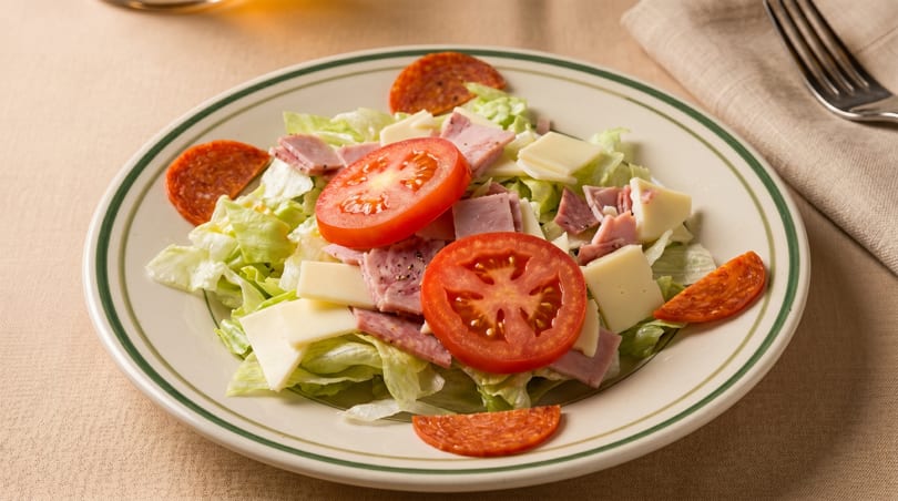 A fresh salad with lettuce, ham, cheese, tomato slices, and pepperoni on a white plate.