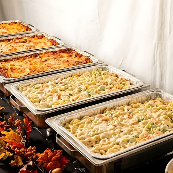 Buffet table with lasagna, pasta salad, and fruit salad in serving dishes, decorated with autumn leaves.