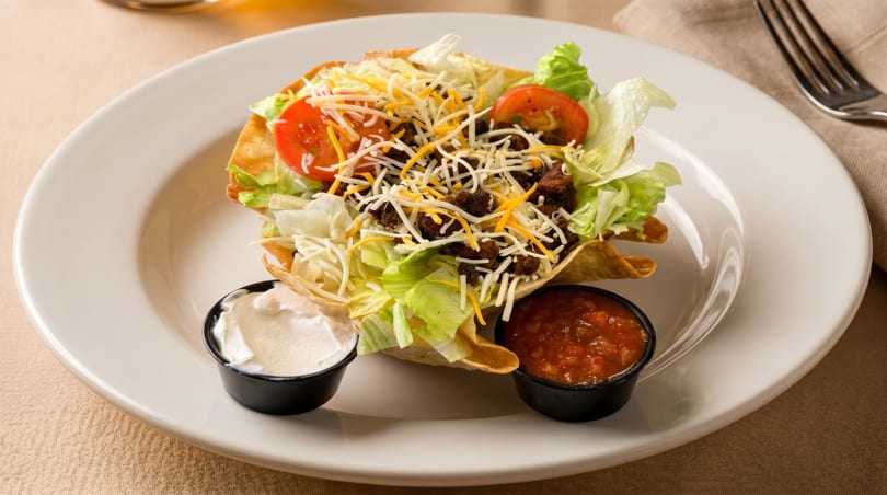 Taco salad in a crispy tortilla bowl with ground beef, lettuce, cheese, tomatoes, and sides of sour cream and salsa.