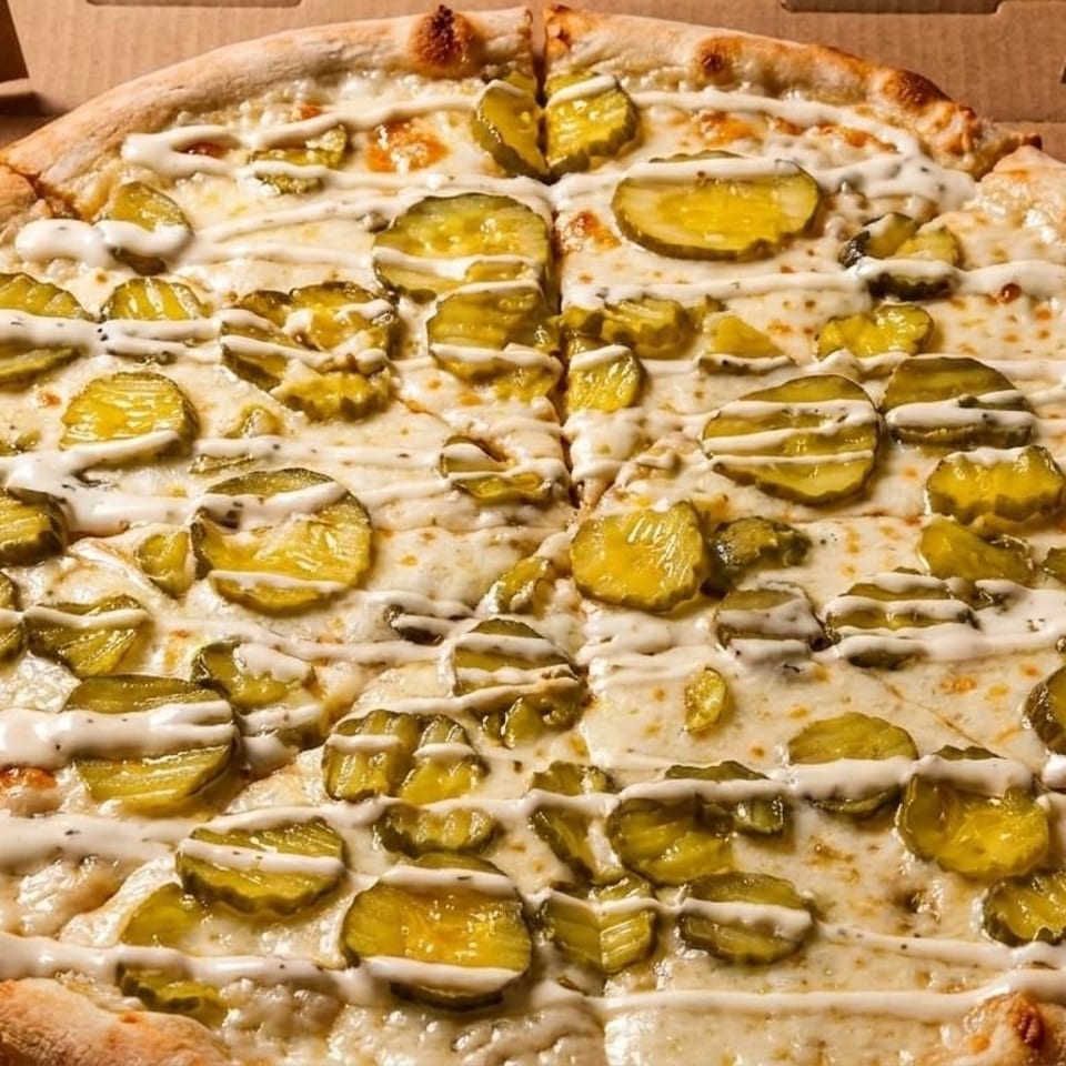 Whole pickle pizza with white sauce drizzle, in a cardboard delivery box.