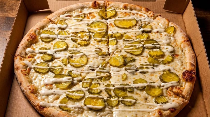 Whole pickle pizza with white sauce drizzle, in a cardboard delivery box.