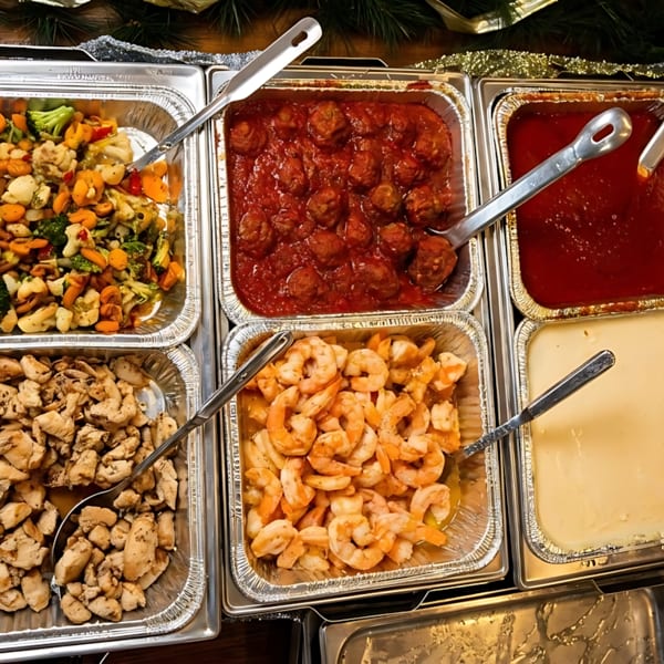 Catering trays filled with meatballs, pasta, chicken, shrimp, vegetables, and sauces.
