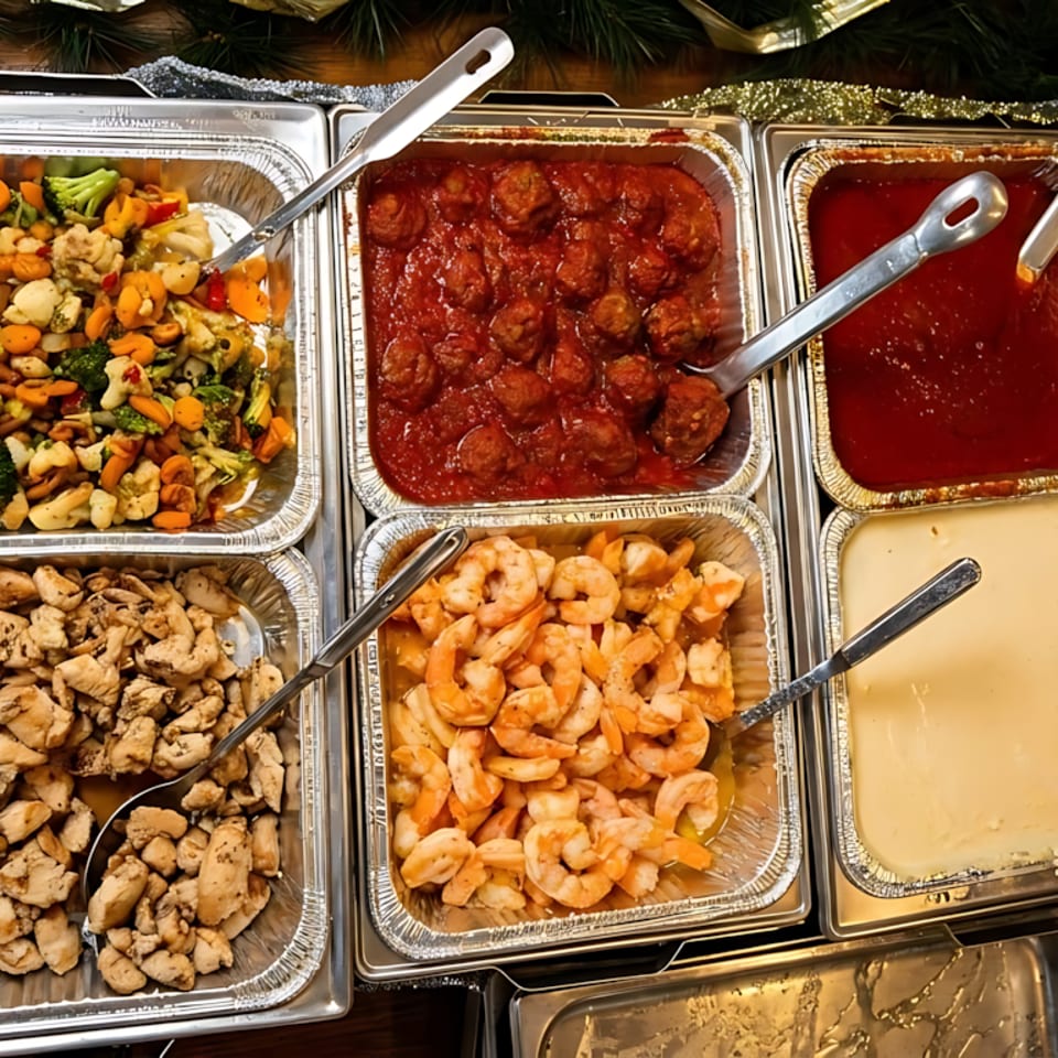 Catering trays filled with meatballs, pasta, chicken, shrimp, vegetables, and sauces.