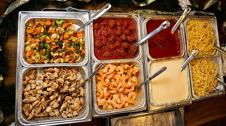 Catering trays filled with meatballs, pasta, chicken, shrimp, vegetables, and sauces.
