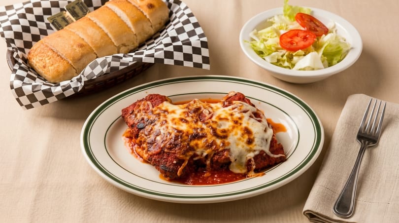 Chicken parmesan with melted cheese, a side salad, and a basket of bread on a tablecloth.