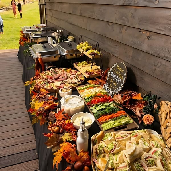 Outdoor buffet table with charcuterie, vegetables, wraps, and chafing dishes, decorated with autumn leaves.