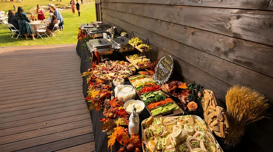 Outdoor buffet table with charcuterie, vegetables, wraps, and chafing dishes, decorated with autumn leaves.